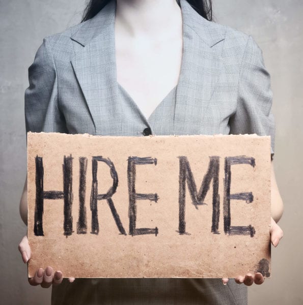 Woman stands behind sign that says "Hire Me"