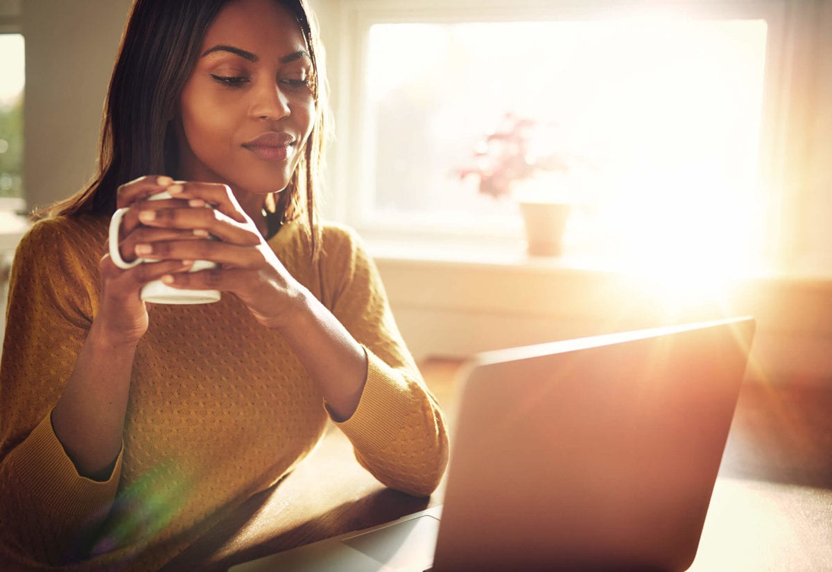 Woman holds coffee mug while looking at computer screen at home