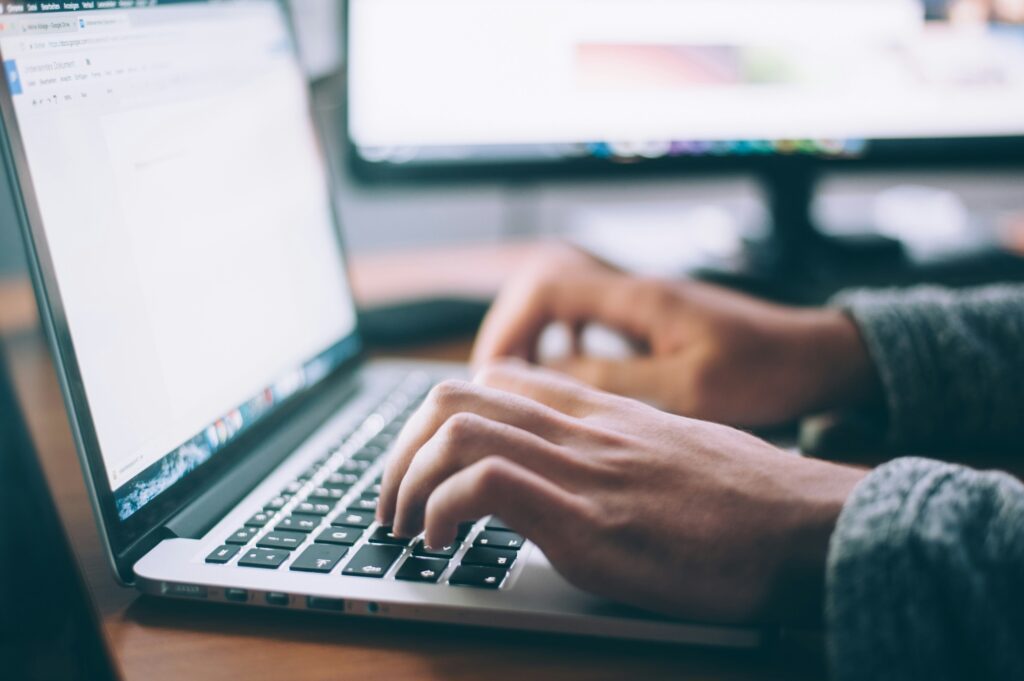 Person typing on a laptop keyboard with dual monitors, illustrating technology use in modern AI talent sourcing.